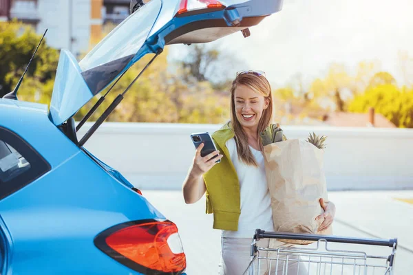 Alışveriş çantalı genç bir kadın arabanın yanında akıllı telefon kullanıyor.