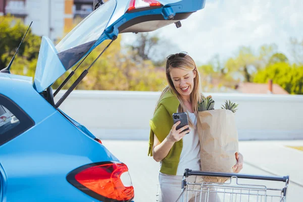 Alışveriş çantalı genç bir kadın arabanın yanında akıllı telefon kullanıyor.