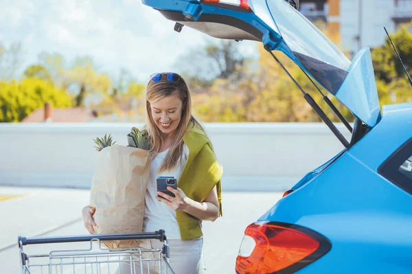 Alışveriş çantalı genç bir kadın arabanın yanında akıllı telefon kullanıyor.