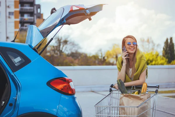 Genç bir kadın arabanın yanında alışveriş arabasıyla dikilirken akıllı telefondan konuşuyor.