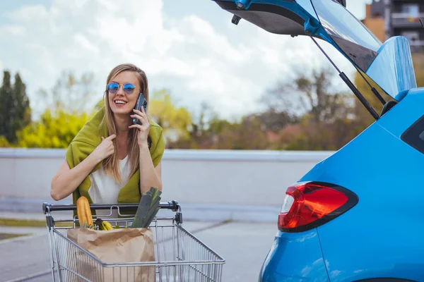 Genç bir kadın arabanın yanında alışveriş arabasıyla dikilirken akıllı telefondan konuşuyor.