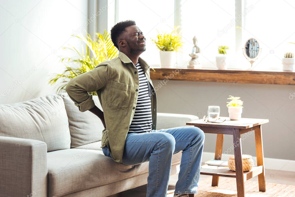 african american man with back pain sitting on sofa 