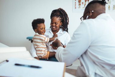 Beyaz üniformalı, gülümseyen bir çocuk doktoru elinde bir not panosu, kontrol toplantısında oğlu olan genç bir anneyi dinliyor, profesyonel çocuk doktoru danışmanlığı, sağlık hizmeti konsepti..