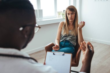 A young woman sits on a couch with her unrecognizable therapist. Psychotherapy session, woman talking to his psychologist in the studio. Psychiatrist listening to her patient who experienced traumatic events, sitting on beige settee