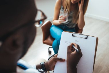 A young woman sits on a couch with her unrecognizable therapist. Psychotherapy session, woman talking to his psychologist in the studio. Psychiatrist listening to her patient who experienced traumatic events, sitting on beige settee