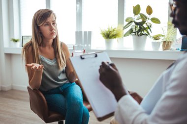 A young woman sits on a couch with her unrecognizable therapist. Psychotherapy session, woman talking to his psychologist in the studio. Psychiatrist listening to her patient who experienced traumatic events, sitting on beige settee