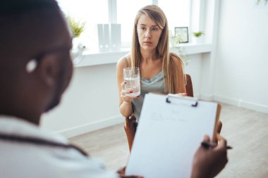A young female adult is feeling sad while at her doctor's office. She has her head down. A young frustrated girl meets with a therapist to discuss her struggles, addictions and mental well being.