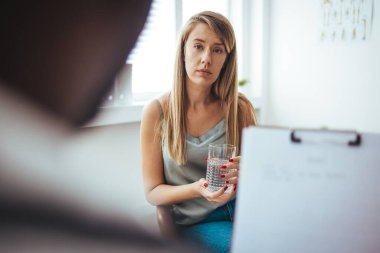A young female adult is feeling sad while at her doctor's office. She has her head down. A young frustrated girl meets with a therapist to discuss her struggles, addictions and mental well being.