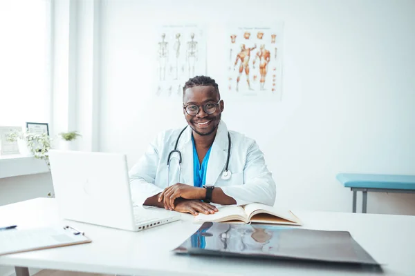 Portrait Of Smiling male Doctor Wearing White Coat With Stethoscope Sitting Behind Desk In Office. Happy male medical doctor portrait in hospital. Portrait of a male doctor with laptop sitting at desk in medical office.