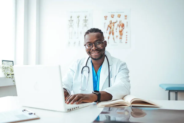 Doctor in office working on laptop. Male doctor working at office desk and smiling at camera, office interior on background. Portrait of doctor in eyeglasses and in white coat sitting at his workplace in front of laptop and looking at camera at offic