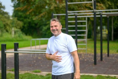 Bir bahar parkındaki spor tesisinde egzersiz barları önünde poz veren mutlu, kıdemli bir adam sağlık ve zindelik konseptinde.