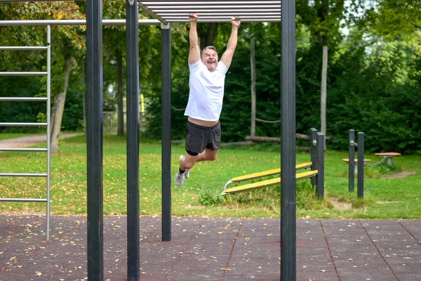 屋外のトレーニングエリアで彼の腕によってバーのセットからぶら下がっ公園で働く楽しみを持っている中年の男 — ストック写真