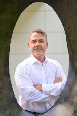 Kendine güveni tam orta yaşlı bir adam, kollarını kavuşturmuş, eski tarz bir grunge tarzı, oval çerçevede poz veriyor ve yoğun bir bakışla kameraya bakıyor.