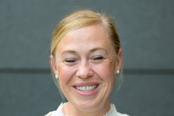 Close up of a happy beaming blond middle-aged woman with closed eyes posing against a grey wall