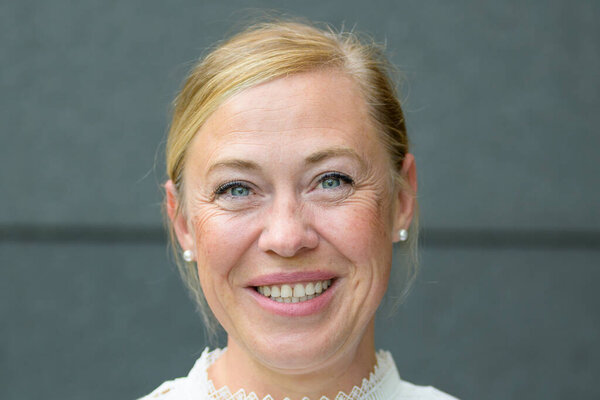 Close up of a happy beaming blond middle-aged woman with opened eyes posing against a grey wall
