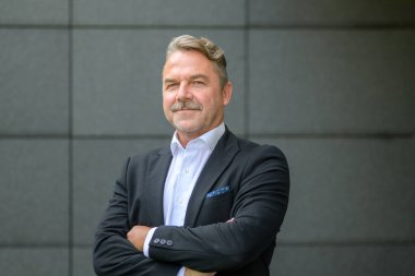 Şık, orta yaşlı, takım elbiseli, kollarını kavuşturmuş kameraya bakan bir adam.