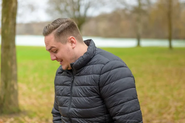 笑顔で見下ろして公園に立って暖かい冬のジャケットで若い男を狙撃 — ストック写真