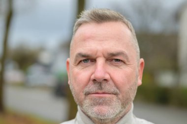 Sakallı yaşlı bir adamın, şehir sokağında ciddi bir şekilde düşünceli bir ifadeyle kameraya baktığı fotoğraf.