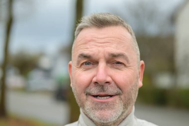 Orta yaşlı, sakallı, açık havada kameraya gülümseyen bir adam.