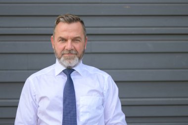 Yoğun orta yaşlı işadamı, gri bir dış duvara karşı ön baş ve omuz portresiyle düşünceli bir şekilde kameraya bakıyor.