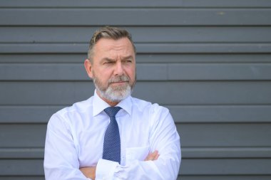 Kafası karışmış kıdemli işadamı, gri bir duvarın önünde, kollarını kavuşturup yakın plan bir portrede poz verirken yüzünü yana çeviriyor.