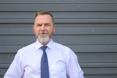 Üst düzey bir adam, gri desenli bir duvarın üzerinde ciddi bir ifadeyle izleyiciyle sohbet ediyor.