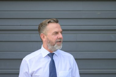 Kafası karışmış kıdemli işadamı, kapalı bir portrede kollarını katlamış gri bir duvarın önünde poz verirken ağzı açık bir şekilde yana bakıyor.