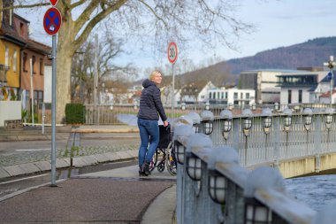 Genç bir kadın kameraya bakarken şehirde boş bir tekerlekli sandalyeyi itiyor.