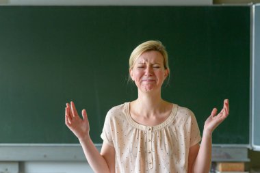Angry frustrated school teacher with her hands up standing in front of a chalkboard in a classroom clipart