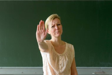 Sinirli ve üzgün bayan öğretmen, temiz yeşil karatahtaya karşı sınıfta duruyor ve avucuyla hareket etmeyi kesiyor, tiksinmiş suratıyla kameraya bakıyor.