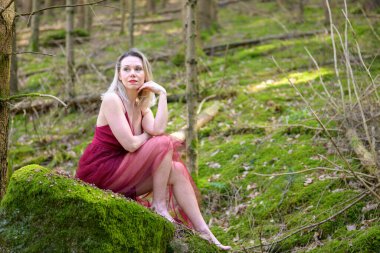 Elleri çenesinde eflatun elbisesi olan düşünceli bir kadın ormanda yosun kaplı kayanın üzerinde oturuyor.