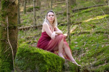 Ormandaki yosun kaplı kayanın üzerinde oturan macenta elbisesi giyen kendine güvenen sarışın kadının tam boy portresi.