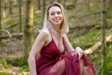 Morumsu bir elbise giymiş, gülümseyen sarışın kadının yan görüntüsü. Ormanda otururken dekolteyi başka tarafa bakıyor.