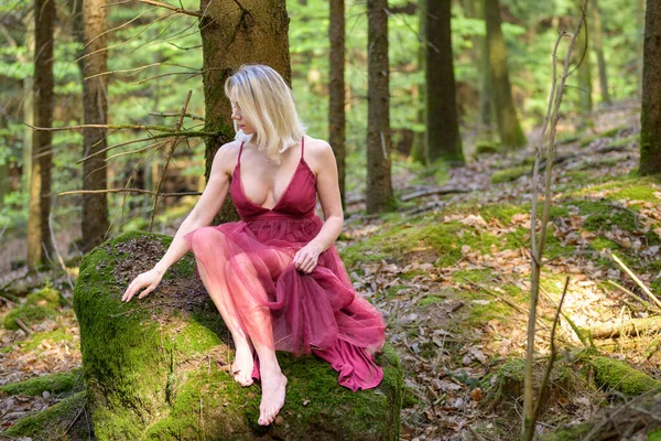Sarışın bir kadın, açık mor elbiseler giyiyor. Göğüs dekoltesi görünüyor. Ormandaki yosun kaplı kayanın üzerinde oturuyor.