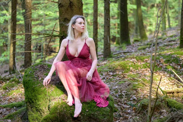 Düşünceli seksi sarışın kadın açık mor elbiseler giyiyor kaya yosunlarının üzerinde oturuyor ve ormanda başka tarafa bakıyor.