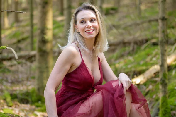 Morumsu bir elbise giymiş, gülümseyen sarışın kadının yan görüntüsü. Ormanda otururken dekolteyi başka tarafa bakıyor.