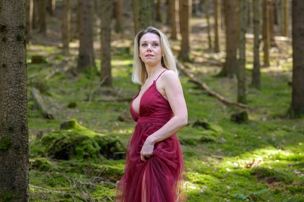 Güzel sarışın kadın macenta elbiseler giyiyor. Ormanda dikilirken gözlerini kaçırıyor.