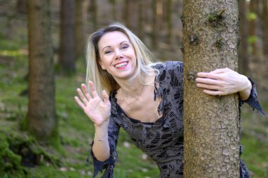 Arsız bir sırıtışla ağacın gövdesine bakan neşeli sarışın kadın kameraya el sallıyor.