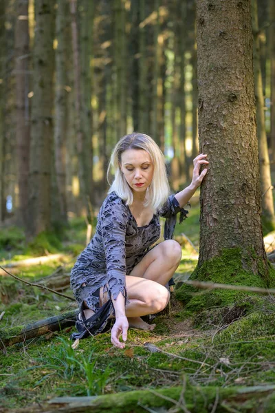 Zarif sarışın kadın, yosunlu orman zeminine uzanırken ormanlık alanda bir ağacın yanında çömelmiş poz veriyor.