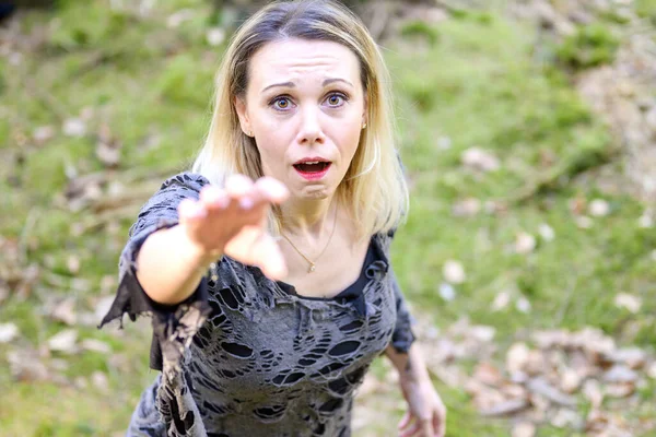 Wild angry blond woman reaching up to claw at the camera with a snarl as she crouches beside a forest tree