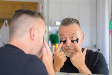 Orta yaşlı bir adam banyodaki aynaya cilt bakımı konseptiyle jel hidrasyon bandı yapıştırıyor.