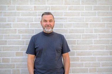 Tuğla duvarın önünde poz veren mavi yazlık gömlekli, sakallı yaşlı bir adamın üst vücut portresi.