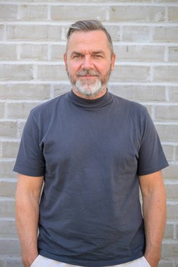 Çekici, orta yaşlı, mavi gömlekli, elleri cebinde, tuğla bir duvarın önünde poz veren bir adam.