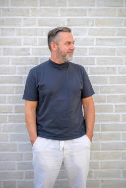 Keyifli, orta yaşlı, rahat bir adam tuğla duvarın önünde dikilip yan tarafa bakıyor.
