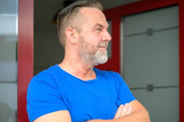 Modern saç kesimi olan sıradan orta yaşlı bir adam kollarını kavuşturup gülümseyerek yana bakıyor.