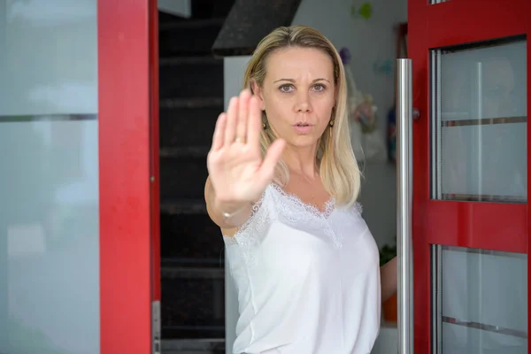 Stern middle aged blond woman holding up her hand in a halt or stop gesture in an open doorway