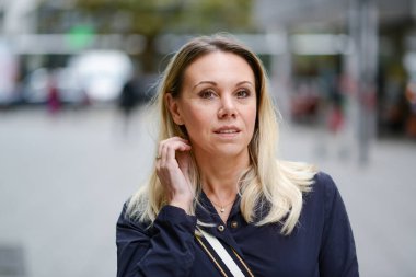 Çekici, orta yaşlı, mavi ceketli sarışın kadın alışveriş caddesinde alışveriş yaparken hafif bir gülümsemeyle kameraya bakıyor.