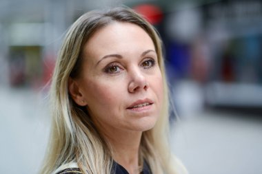 Çekici, orta yaşlı, mavi ceketli sarışın kadın sakin bir bakışla bir alışveriş merkezinin arka planına bakıyor.