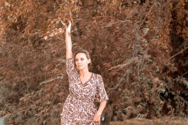 Orta yaşlı sarışın kadın, sonbahar ağaçlarının önündeki kameraya şehvetli bir bakışla uzanıyor.