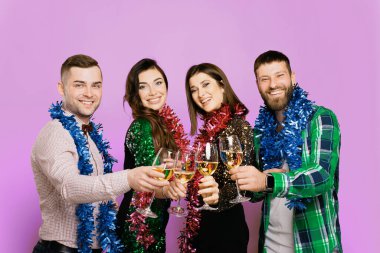 Two men and two girls 25s with New Year's rain on their shoulders clink glasses of white wine or champagne against a lilac background. Four friends with glasses of wine. New Year's party of friends.